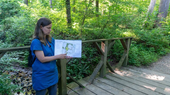 一名女子站在一条绿树环绕的徒步小径上的一座桥上,指着亚马逊HQ2的蓝图。