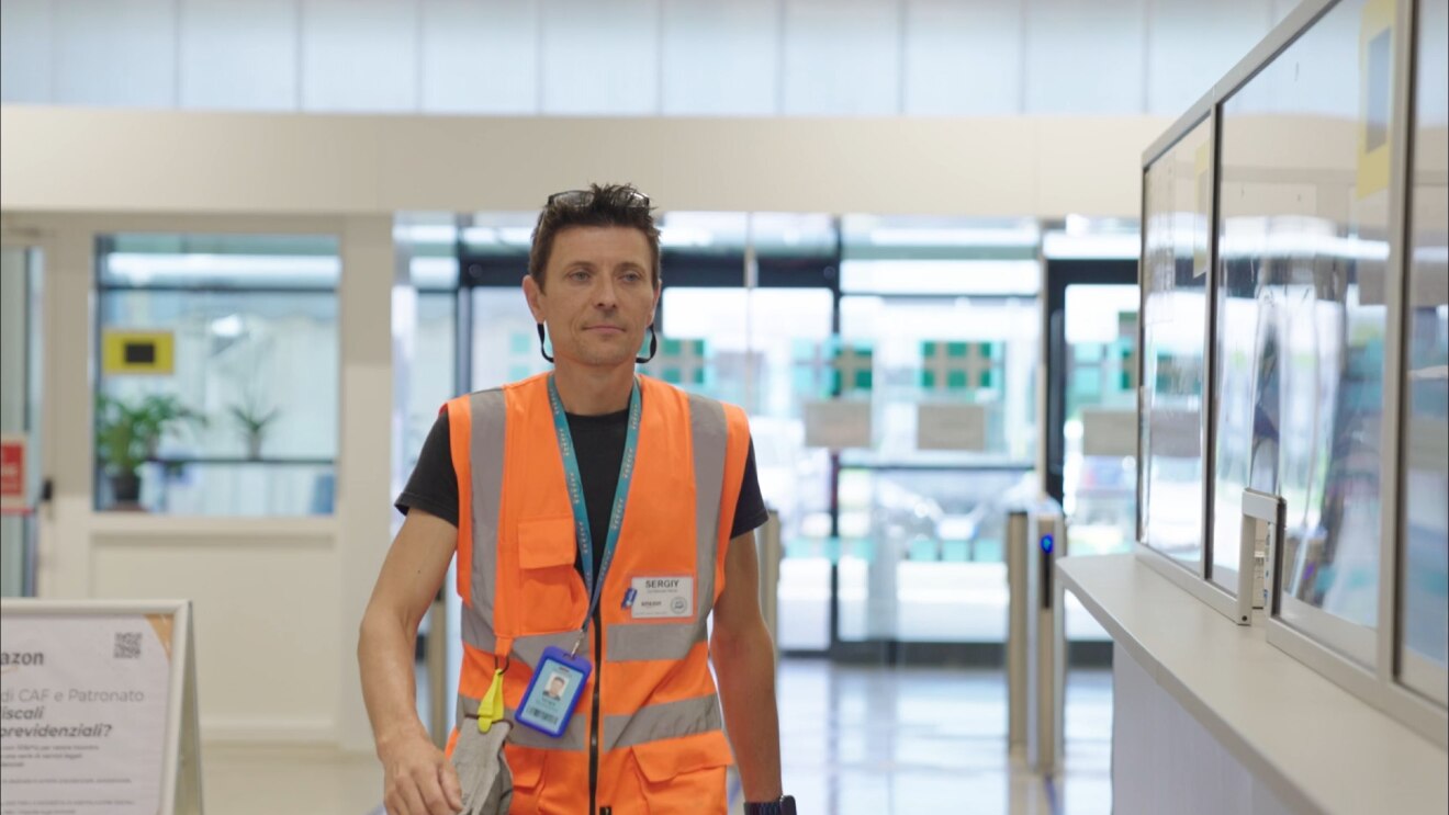 Mezzo busto di un dipendente Amazon che sorride verso la camera. Indossa un giubbino di sicurezza arancione