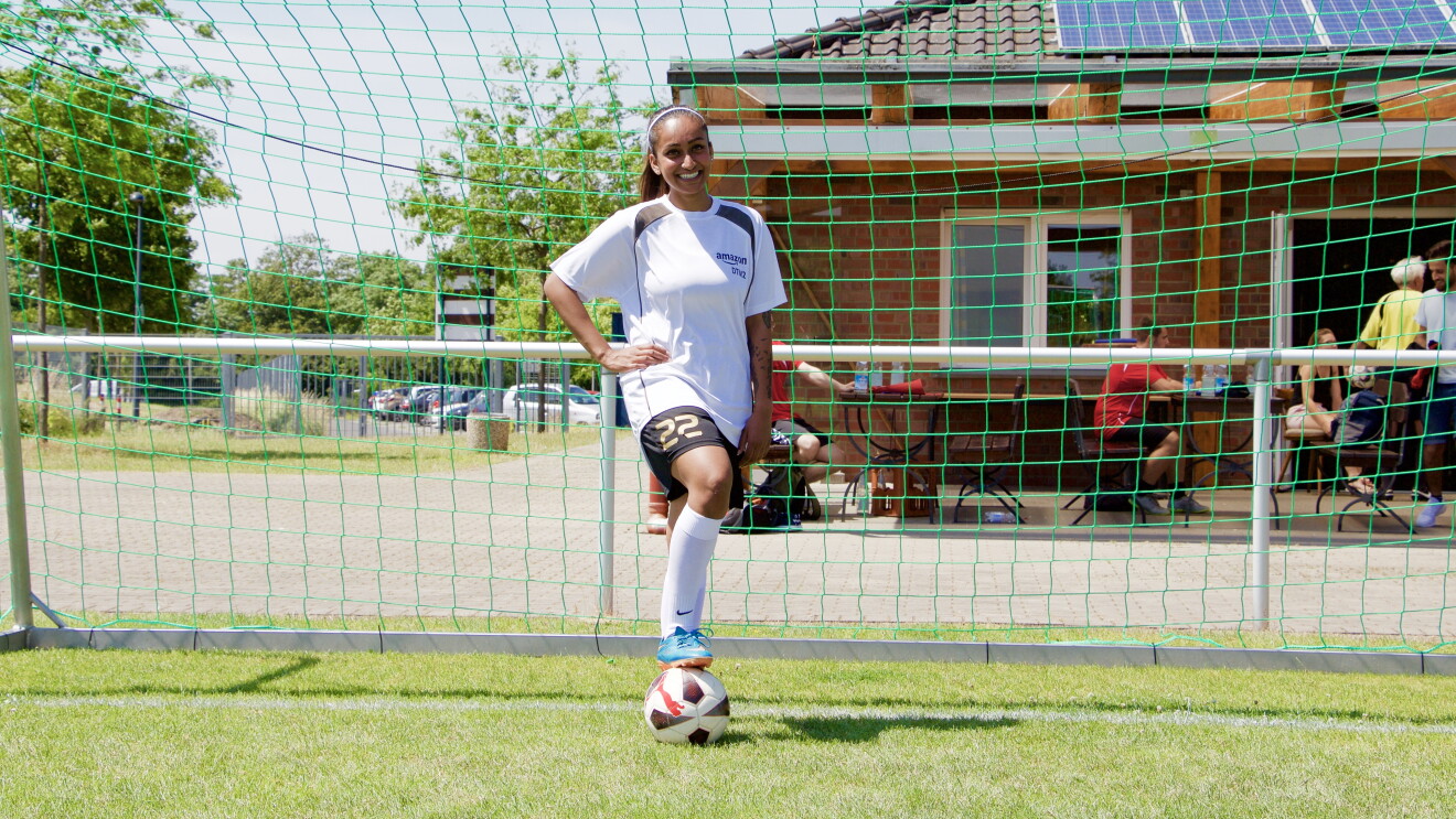 Eine dunkelhäutige Fußballerin vor einem Tor: Sie trägt ein weißes Sportshirt, ihr Fuß ist locker auf einem Ball abgelegt.