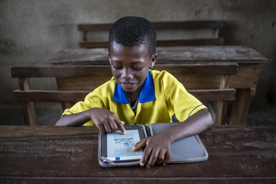 A young boy in a yellow and blue school uniform reads from his Kindle on top of a battered wooden desk.