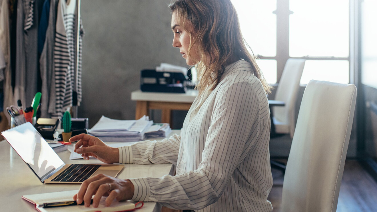 Blonde Frau sitzt am Tisch und arbeitet am Laptop. Im Hintergrund sieht man Stoffe und Kleiderstangen.
