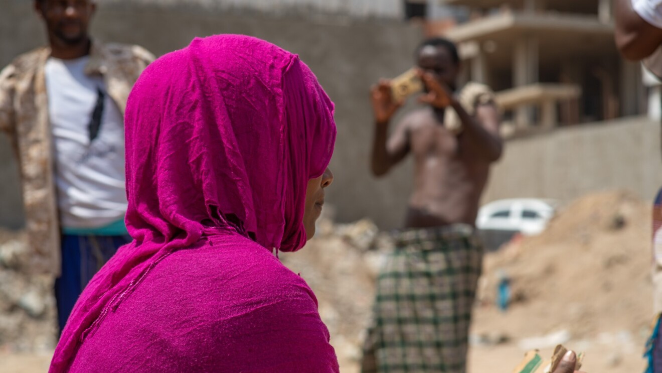 A migrant woman wears a bright pink head scarf, looking away from the camera while sitting in a large open space. Fellow migrant men are also around her in the area.