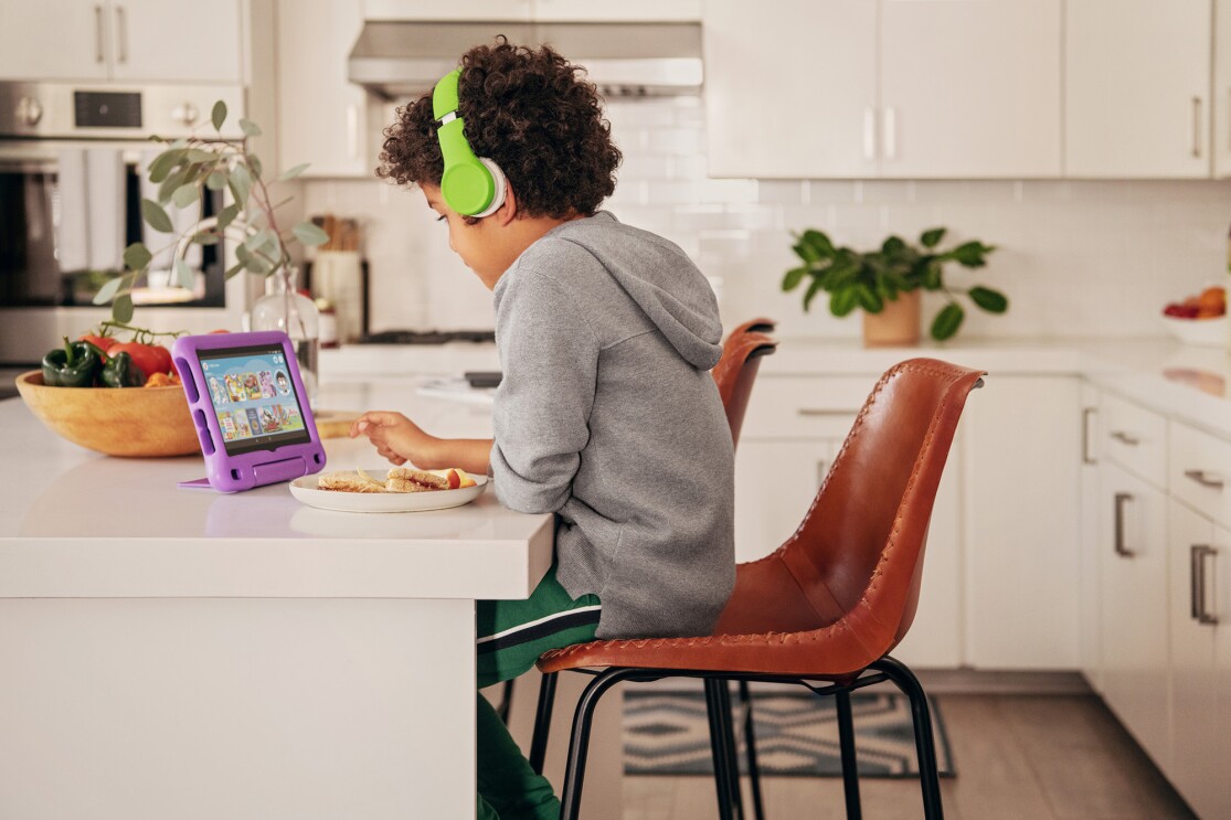 A child sits at a kitchen island, looking at a fire tablet. He wears headphones as he uses the device. 