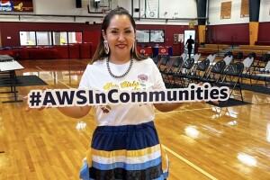 A photo of a member of the Confederated Tribes of Umatilla Indian Reservation standing in a gym, holding a sign that states, #AWSInCommunities.