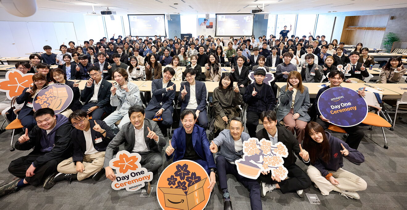 広いホールに集まり集合写真を撮る多数の人々。最前列の7人は床に座っている。2列目は椅子に座り、3列目以降は長机に向かって着席している。