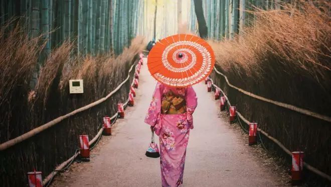 ピンク色の着物を着ている人が赤い傘を持って竹林の小径に立っている壁紙。その一部にエアコンのコントロールパネルが見える