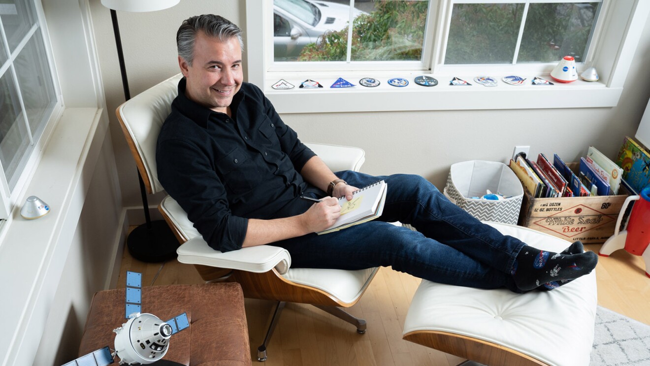 An image of a man sitting in a chair in his office. He is looking up at the camera and smiling while drawing on a notepad and there is a small satellite device on a table next to him.