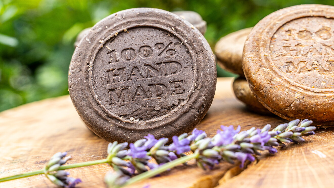 La cosmética de María ofrece dieciocho productos.  En la foto hay dos jabones sólidos, con  una ramita de lavanda y encima de un tronco. Los jabones son de color redondo y tienen grabado lo siguiente: 100% handmade. 