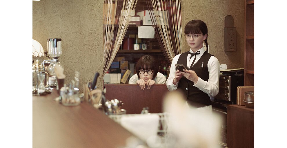 喫茶店のカウンターの中、ウェイトレス姿の女性がスマホを持って立っている。その奥に、仕切りの上から顔をのぞかせる別な女性。