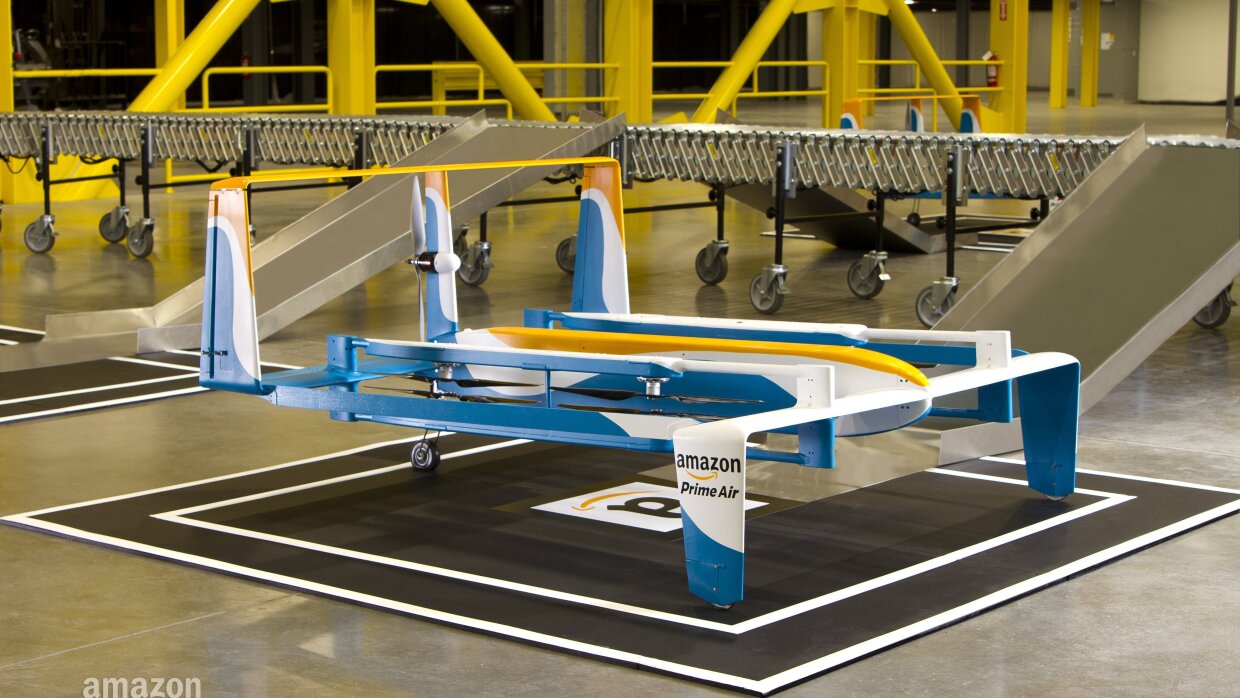 Primer plano del dron de Amazon ubicado en el interior de un centro logístico. El dron es de color blanco, azul y amarillo.