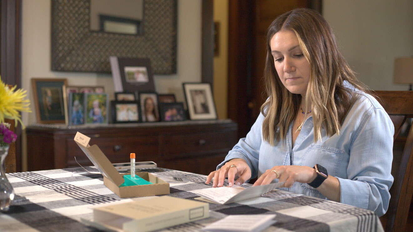 An image of a woman sitting at her kitchen counter with an at-home COVID test in front of her. She is reading directions that came with the test.