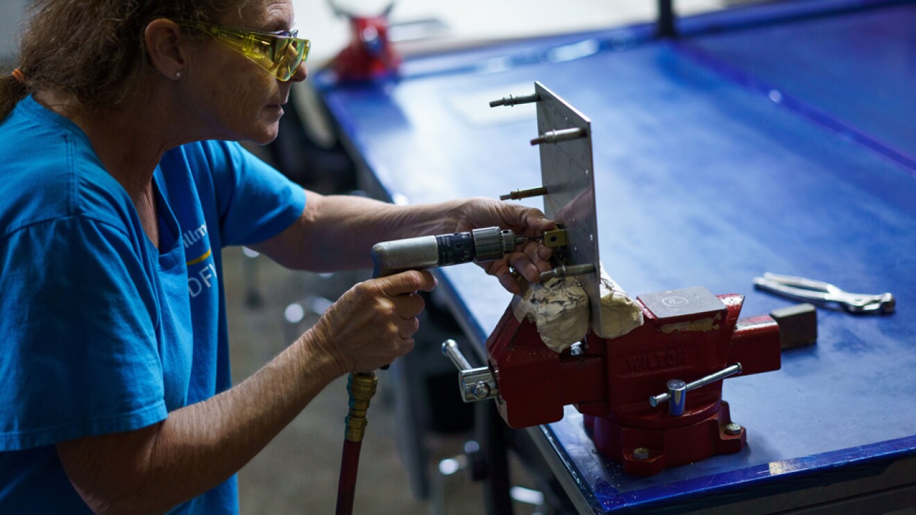 一个穿蓝色t恤的女人拿着工具,靠向工作台上的虎钳。