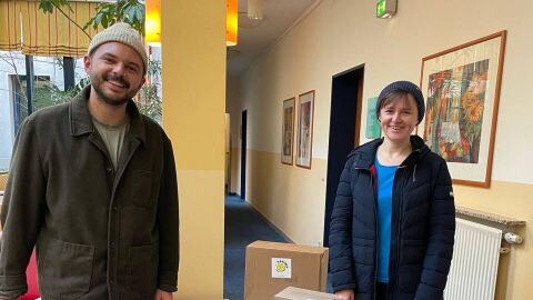 Zwei freiwillige Helfer von Amazon stehen neben diversen Kisten mit Sachspenden wie Powerbanks, Lebensmitteln und Babyartikeln für die gemeinnützige Organisation KARUNA eG.