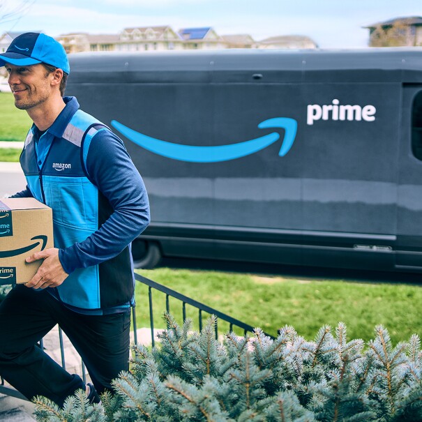 Amazon Prime delivery driver walks up the steps to deliver a package with his Prime delivery van in the background.