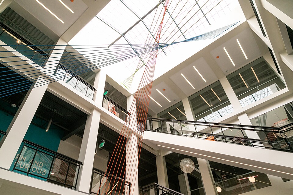 The inside of an office building, with glass ceiling to let light into the space, exposed ceiling spaces, glass-panel lined walkways. An art installation made of strings in orange, leaf green and blue take up the center space of the area.