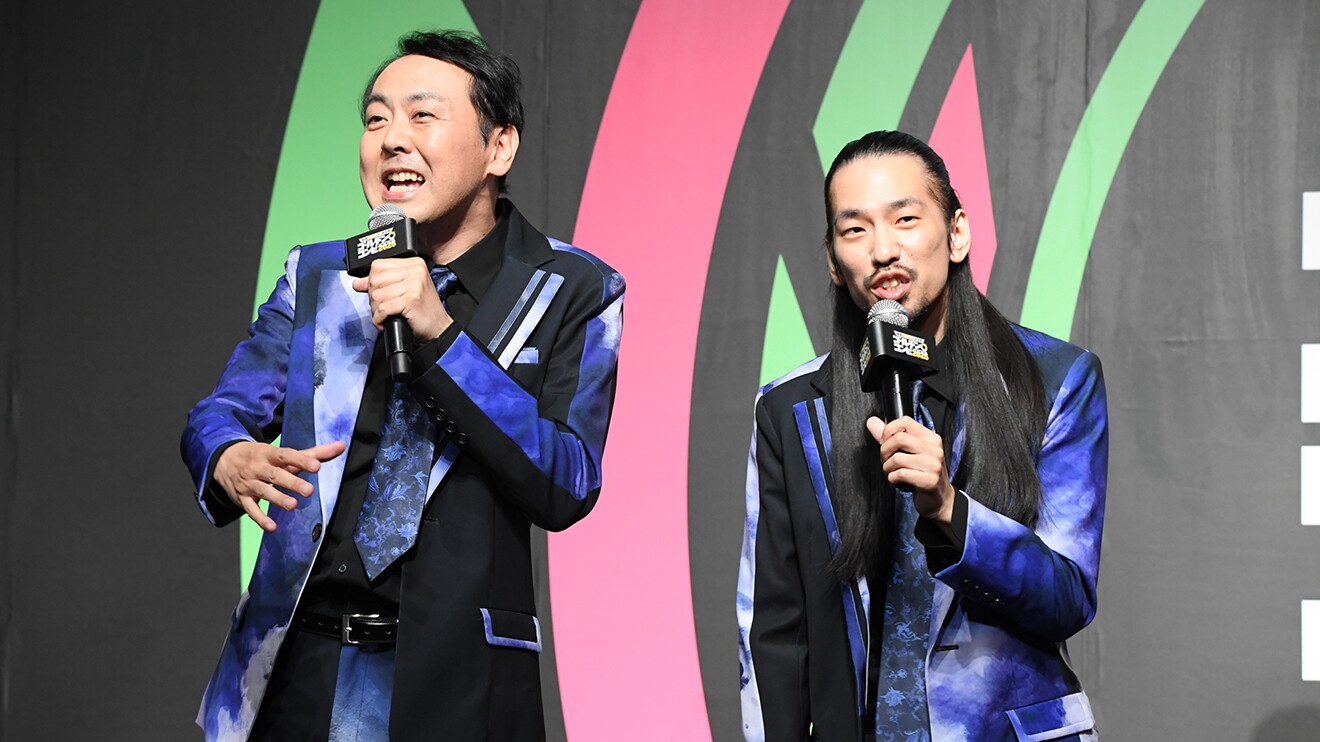 黒地にピンクと黄緑の曲線が入った背景の前で、黒と紺色のマーブル模様のおそろいのスーツを着た2人の男性がマイクを持って立っている。