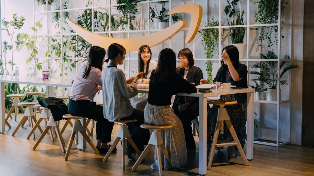 明るい光が差し込むAmazonのロゴが飾られているスペースで、テーブルを囲み談笑している6人の女性たち