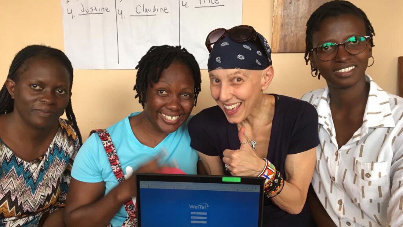 An image of four women smiling for a photo. There is a laptop in front of them and on the screen is the login screen for "WeTel"