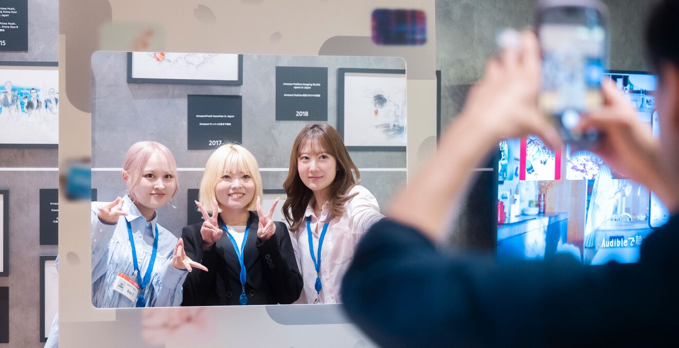 四角くくりぬかれたボードの背後に立ち、記念写真を撮る3人の女性。首から青いひもの社員証を下げている。右側手前に、スマホを構えて撮影する人。