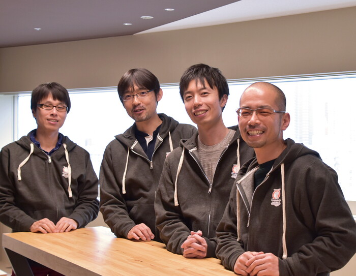 広い窓を背に、そろいのグレーのパーカーを着て並ぶ4人の男性。