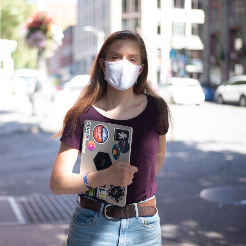 A woman stands on the street with a mask on, looking at the camera. She's holding a laptop with stickers related to Amazon and AWS.