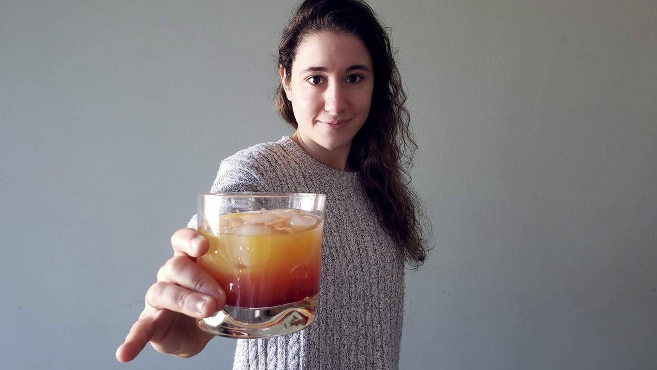 An image of a woman holding a cocktail glass out toward the camera while softly smiling.