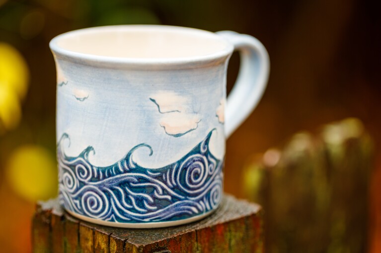 Ceramic mug with painted waves and clouds sits on a wood display pillar.