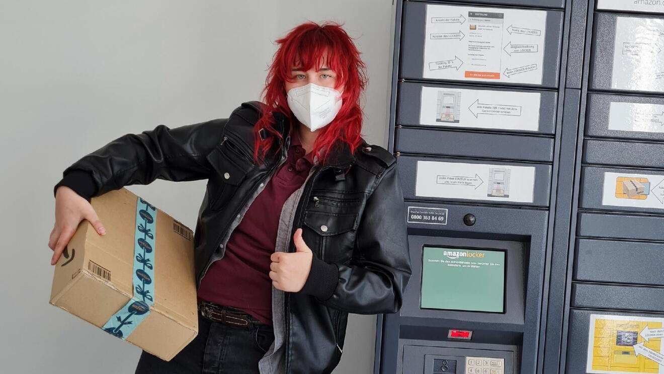 eine Frau mit roten Haaren, Lederjacke und Maske vor einem Amazon Locker: Sie hat ein Amazon Päckchen in der Hand.