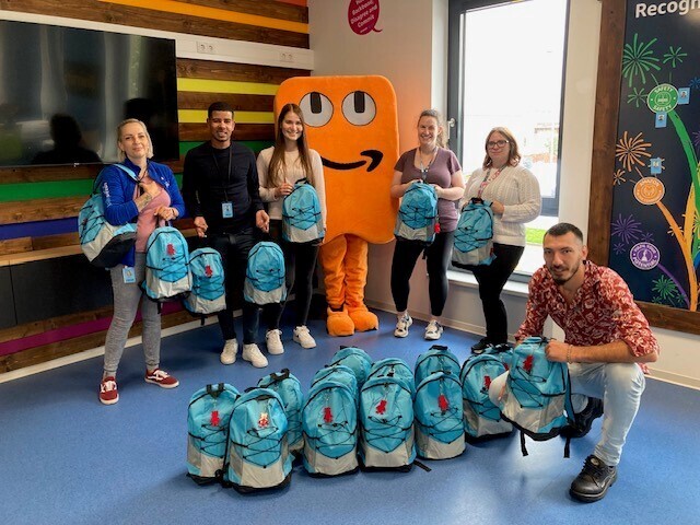 Amazon Mitarbeiterinnen und Mitarbeiter in Kaiserslautern und Maskottchen Peccy stehen vor bunten Back to School Rucksäcken