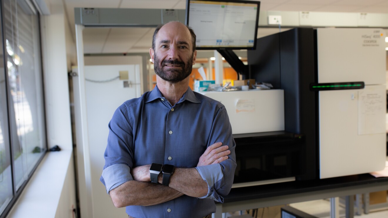 Michael Snyder, PhD, professor and chair of genetics for Stanford University’s School of Medicine is wearing a blue button up with his sleeves rolled up and stands with his arm crossed wearing two smart watches. He stands in his labs with white boards and monitors behind him.