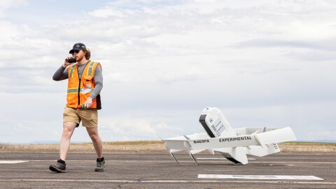 身穿橙色安全背心的亚马逊员工站在亚马逊无人机旁的图片。无人机在空中飞行的图片。