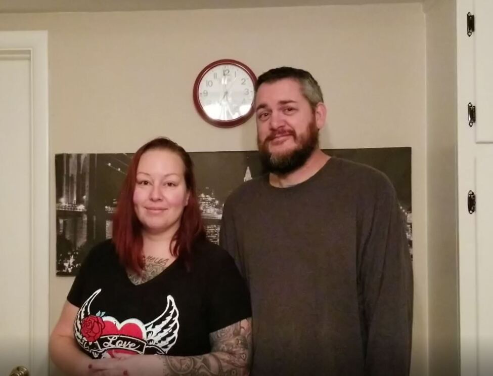 A woman stands next to a man. A clock hangs behind them, on the wall.