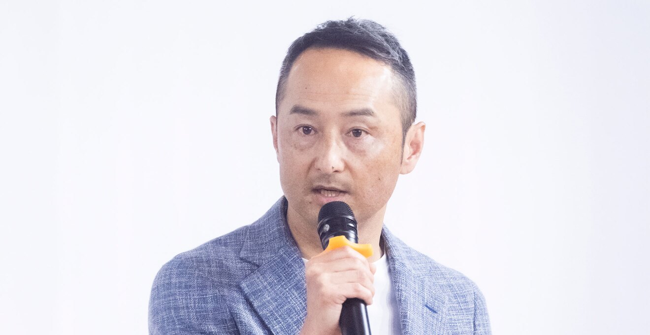 白い壁の前で、マイクを手に話しをする男性。絣の模様が入ったブルーのスーツを着用、インナーは白い丸襟シャツ。