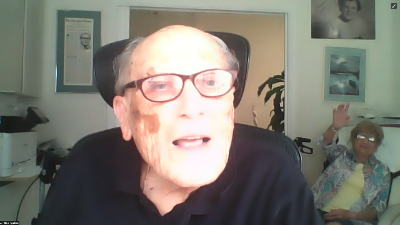 An image of a man on a video conference call. His wife sits in the background smiling and waving at the camera.