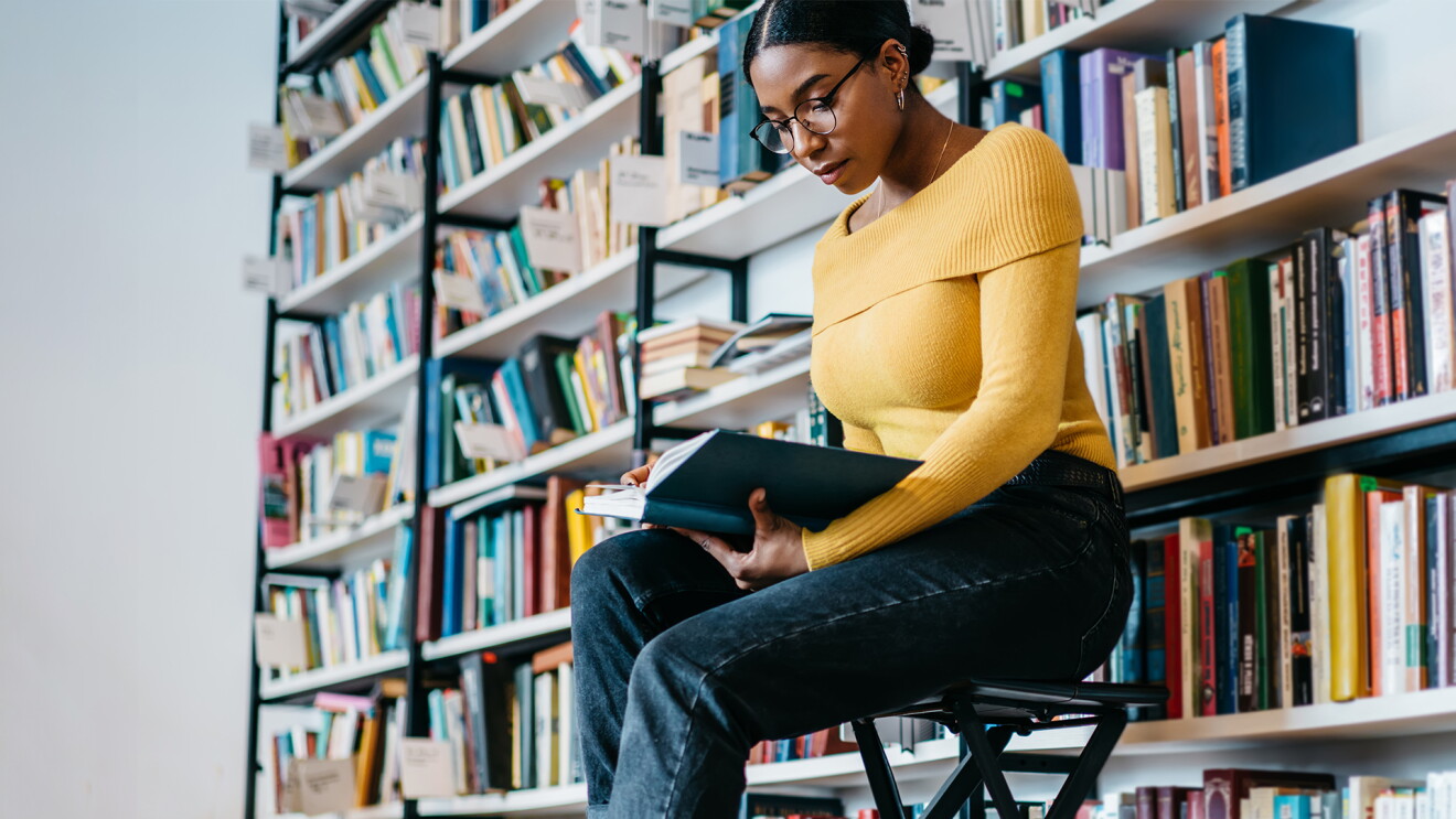 A Black woman wears a yellow sweater and reads a book as she sits in front of bookcases filled with books.