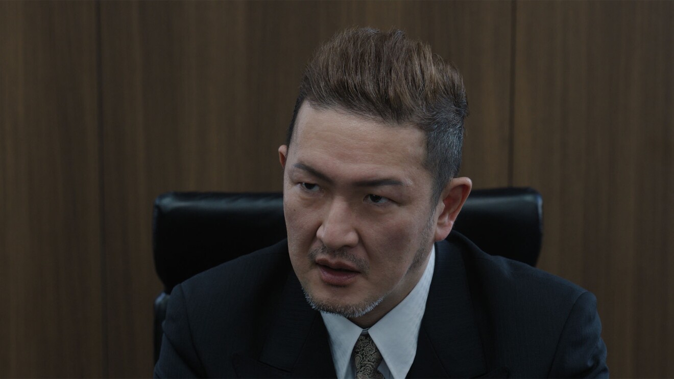 木製の壁の前に置かれた黒い革のハイチェアに座るダークスーツを着た茶髪の男(中村獅童)が険しい表情で話している