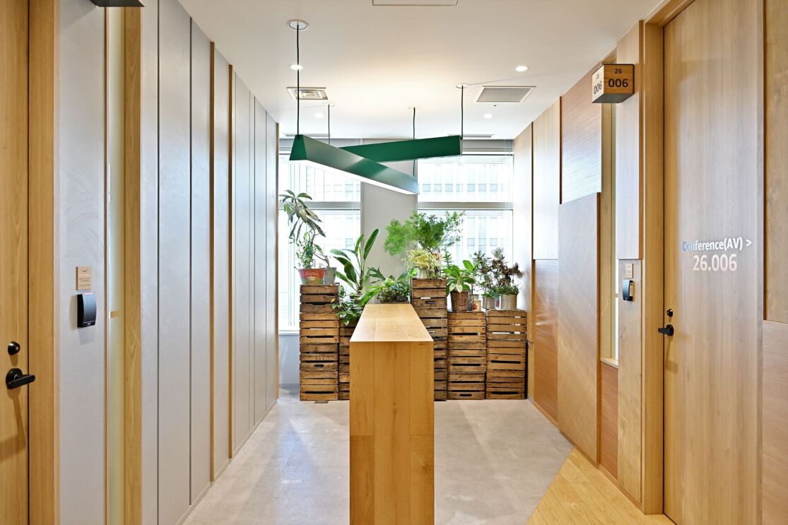 木材で作られたホールに会議室が囲まれている。そのホールの後ろには木箱の上に鉢植えが置かれている。