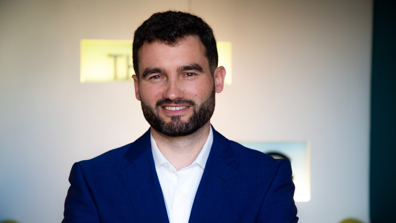 Antonio González, Profesor de venta y marketing digital en IE Universit. Aparece Antonio en un plano corto por los hombros. El pelo negro corto con barba negra de cuatro días. Americana azul, camisa blanca. De fondo pared blanca y una letras iluminadas que no se pueden leer porque están difuminadas. 