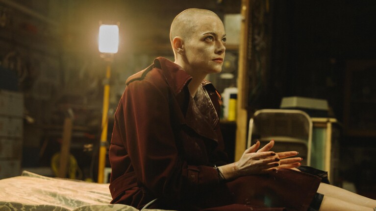 Bald figure in red coat gazes intently in dimly lit, industrial setting
