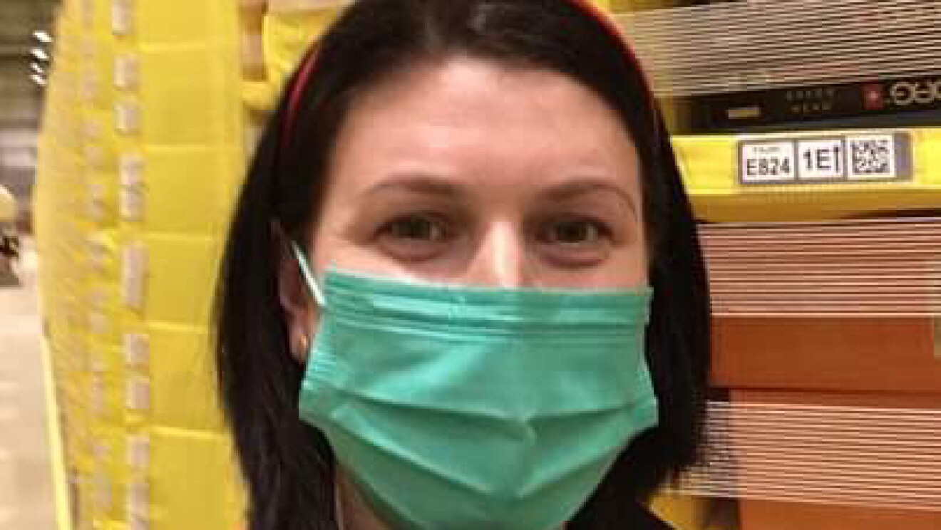 Diana Campean, Associate at Amazon's fulfilment centre in Dunstable pictured in front of lots of yellow pods full of inventory. Diana is wearing a green face mask and a festive headband with two snowmen.