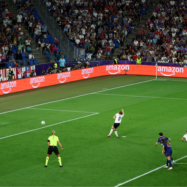 Womens football photo showing a players on the pitch with orange, Amazon branding at the edge of the pitch