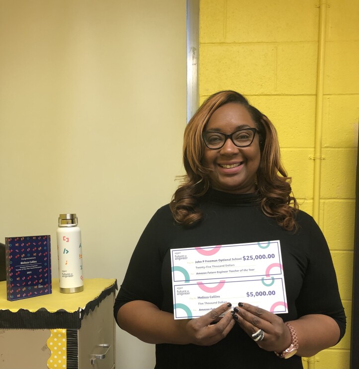 A woman smiles for a photo while holding two checks she earned as an award a recipient of Amazon Future Engineer's Teach of the Year award. One check is for $25,000 and the other is for $5,000.