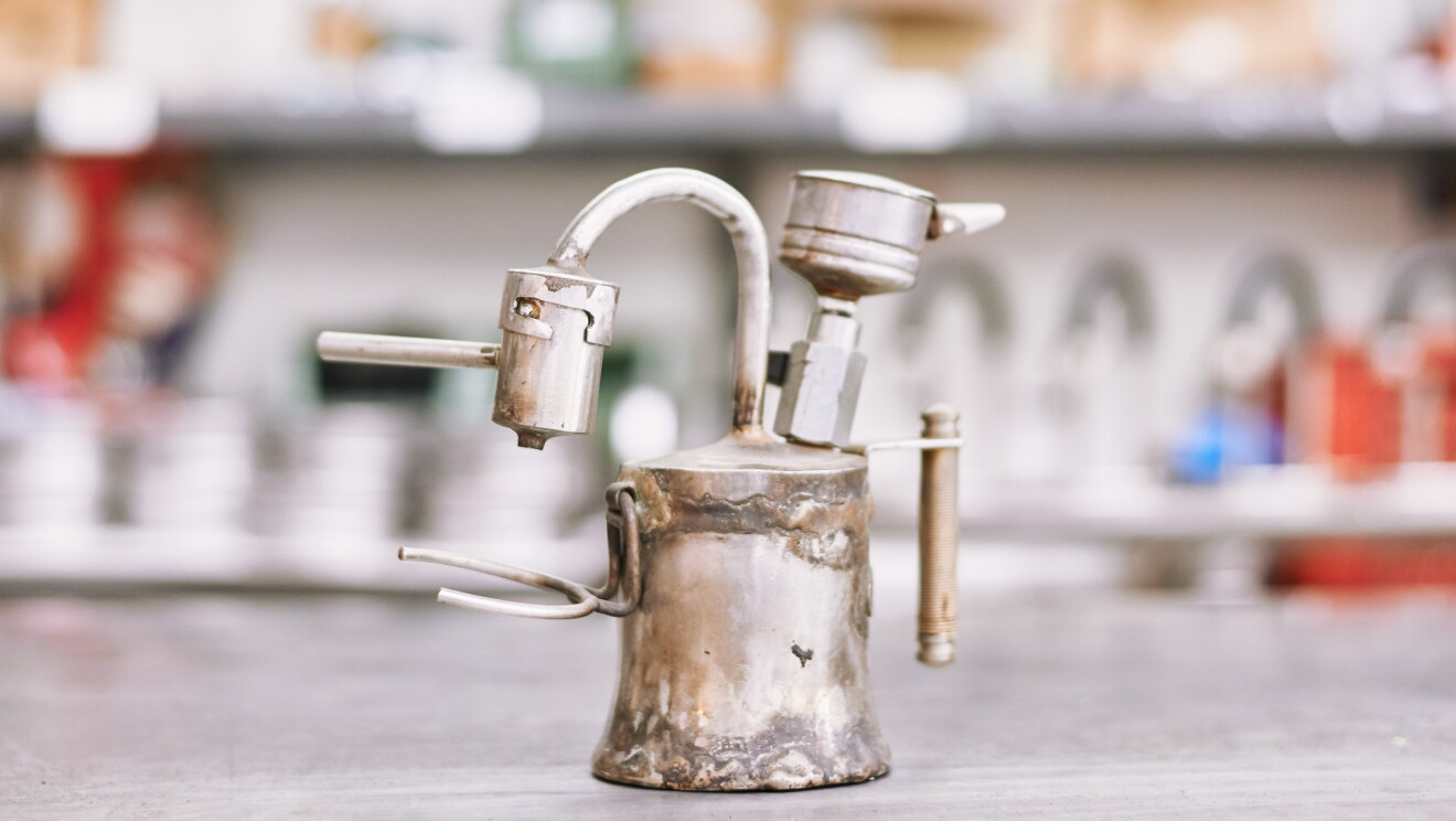 Foto di un prototipo vecchio e piuttosto malandato di caffettiera Kamira, composto da pezzi recuperati da altre caffettiere. Una forcella è attaccata al corpo della caffettiera per reggere la tazzina. Sono molto evidenti le saldature fatte a mano - che questo modello vecchiotto sia esploso durante i test?