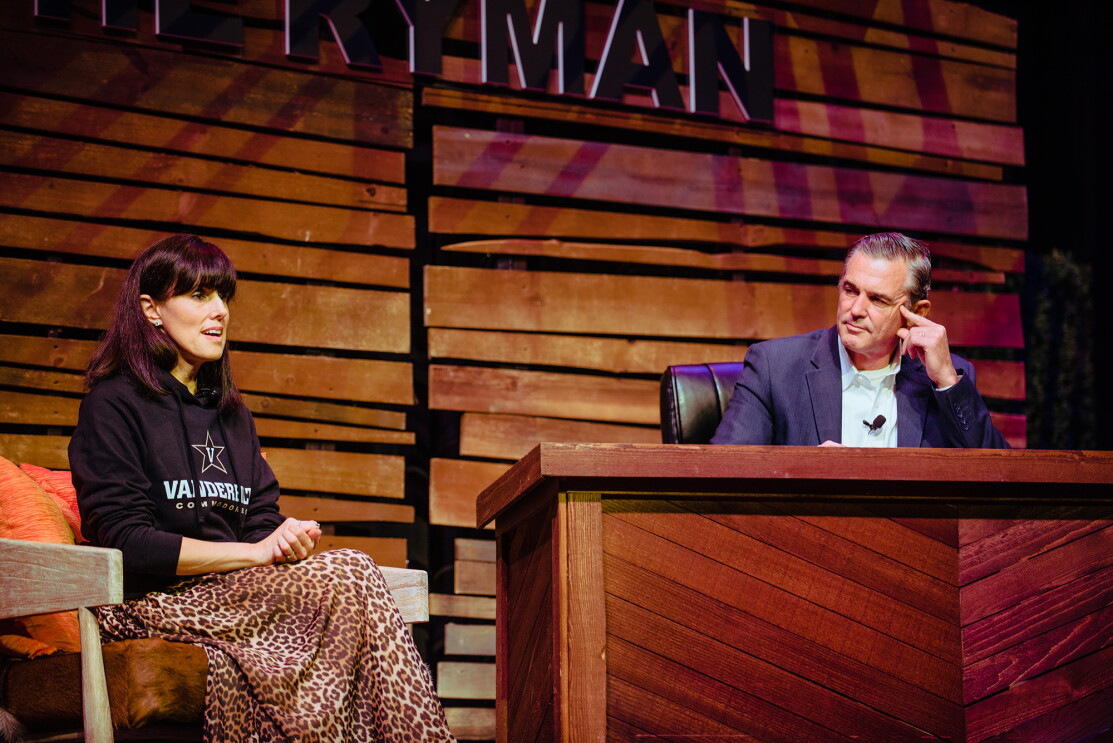 Maria Renz of Amazon wears a Vanderbilt sweatshirt and animal print skirt. She's seated on a stage at The Ryman, next to a desk, with Kevin Omarah seated behind it. They engage in conversation.