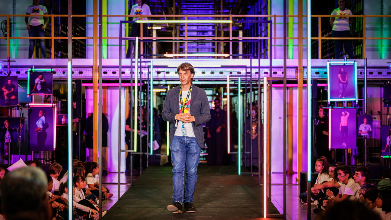 Dentro de un centro logístico, con las estanterías de fondo y las barandillas de color amarillo, aparece una pasarela de color negro ambientada con luces de neón. Nacho Alonso, general manager de MAD4 caminando por la pasarela. Lleva una americana, camiseta del evento blanca coon pinceladas de colores, jeans y zapatos azules. Manos cruzadas delante, badge y pelo castaño.