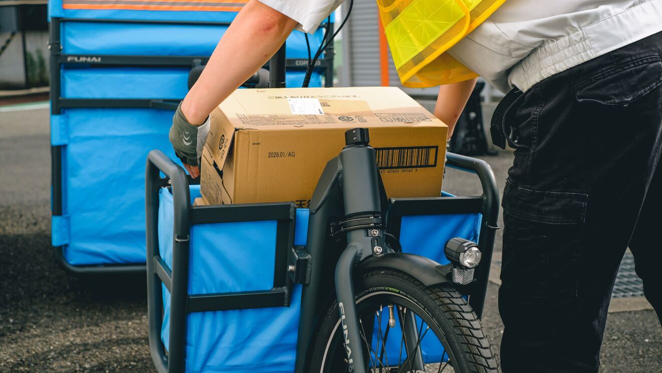 黄色い安全ベストを着ている人が大きなダンボール箱を自転車の前輪部分の青いかごに入れている