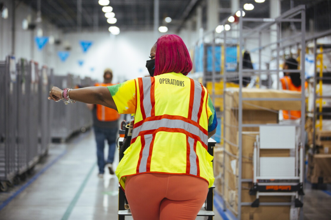 Wearing a yellow Operations safety vest, Auhzha Wright walks away from the camera and down an aisle at Amazon's Fulfillment center with her left arm extended and pointing out of frame.