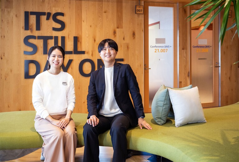 モスグリーンの大きなソファに2名が着席している。右側の人物はAWSロゴ入りの白いスウェットとピンクベージュのパンツを着用。左側の人物はネイビーのジャケットセットアップ。人物の右側に3つのクッション。背後の壁面は木製で、「IT'S STILL DAY ONE」の黒い文字で装飾されている。右奥には会議室へと続く2つのドアと観葉植物が見える。
