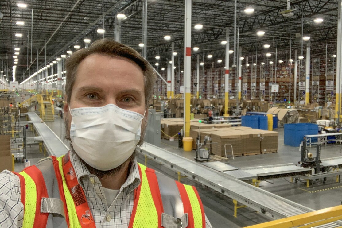 A man wearing a protective mask and a safety vest stands in front of a large warehouse space.