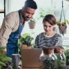 A man stands next to a seated woman, as they look at a computer monitor. Their surroundings suggest they work in a small business that sells plants. 